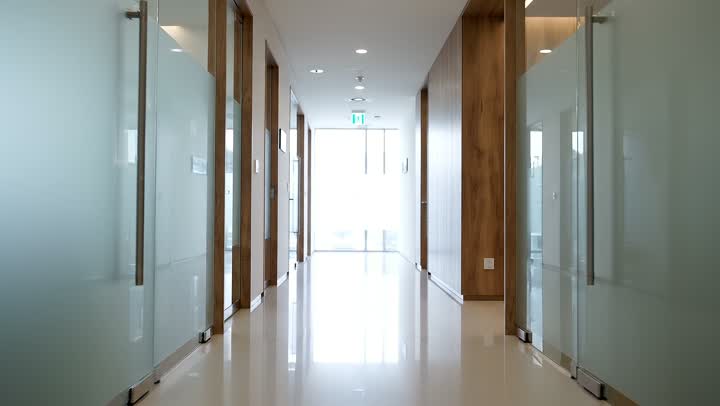 A bright modern clinic corridor with frosted glass doors and a polished floor, looking toward daylight at the far end
