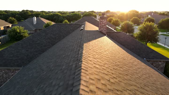 A shingle roof on a suburban house at golden hour, ridge vents and a brick chimney, seen from just above the ridge line